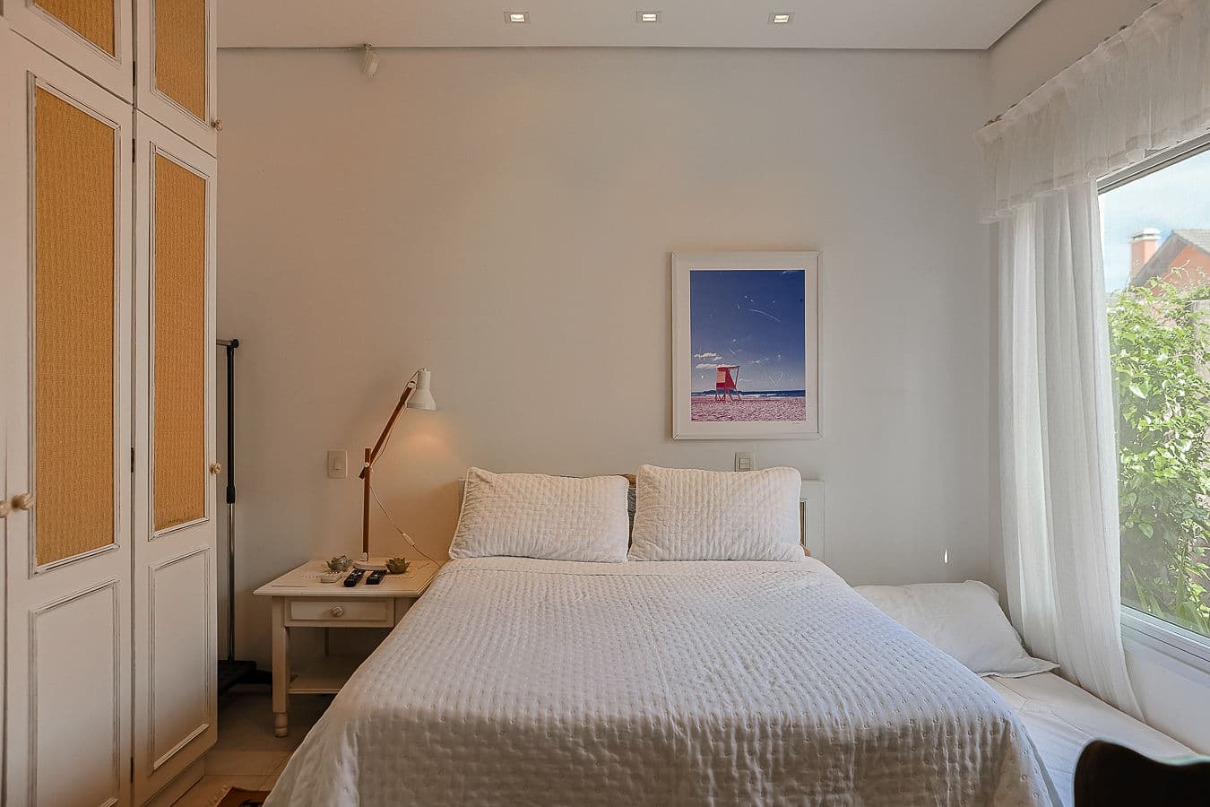 Quarto em tons claros com quadro de praia e luz natural