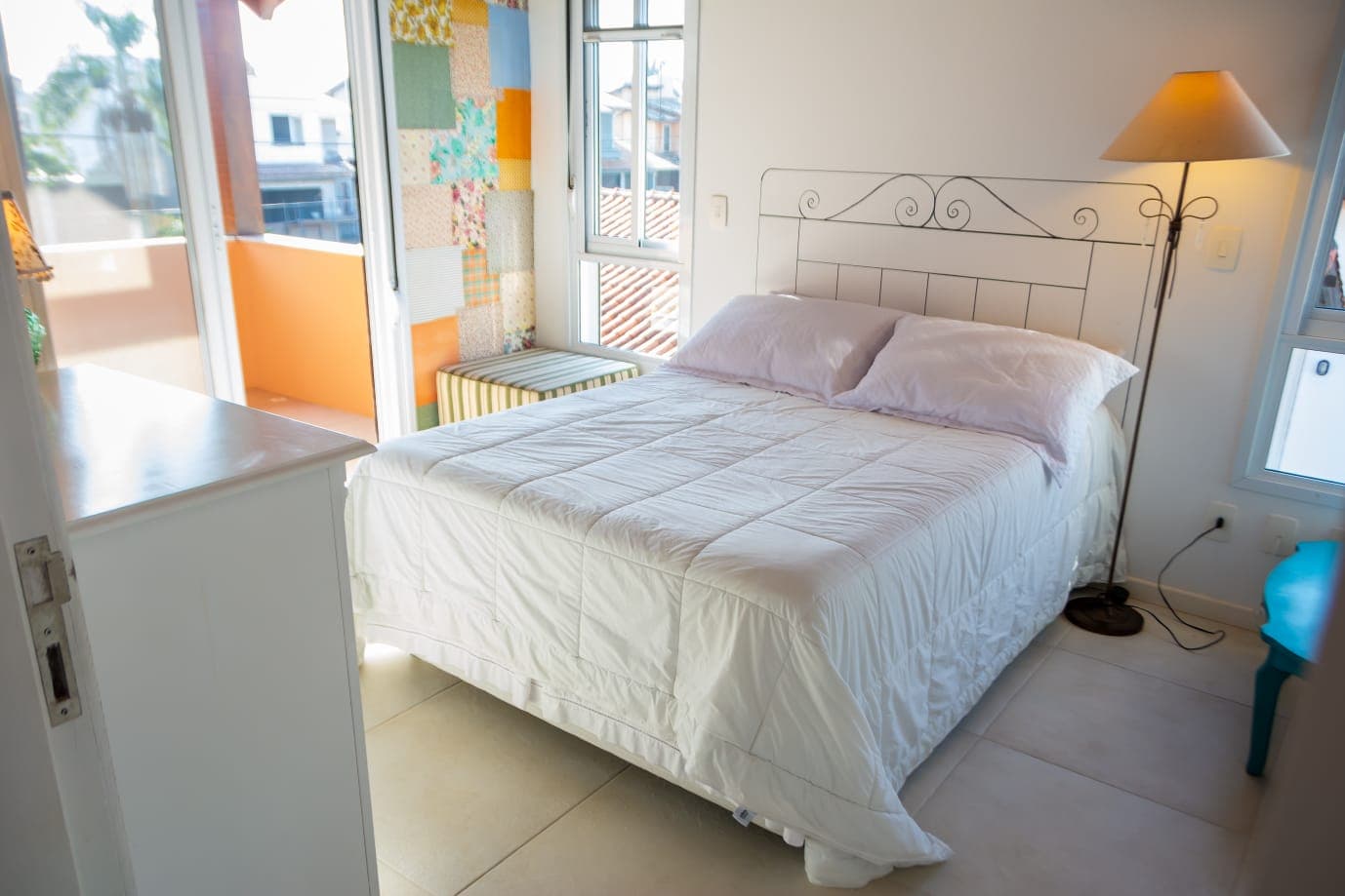 Suíte clara e iluminada com cama de casal, abajur e acesso à varanda