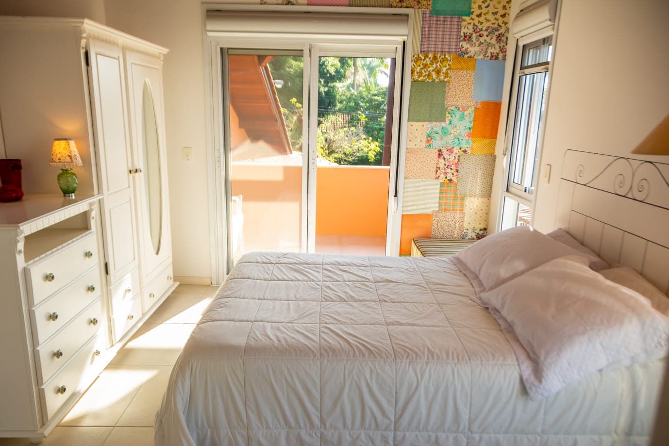 Suíte ensolarada com guarda-roupa, cômoda e porta para a varanda com vista